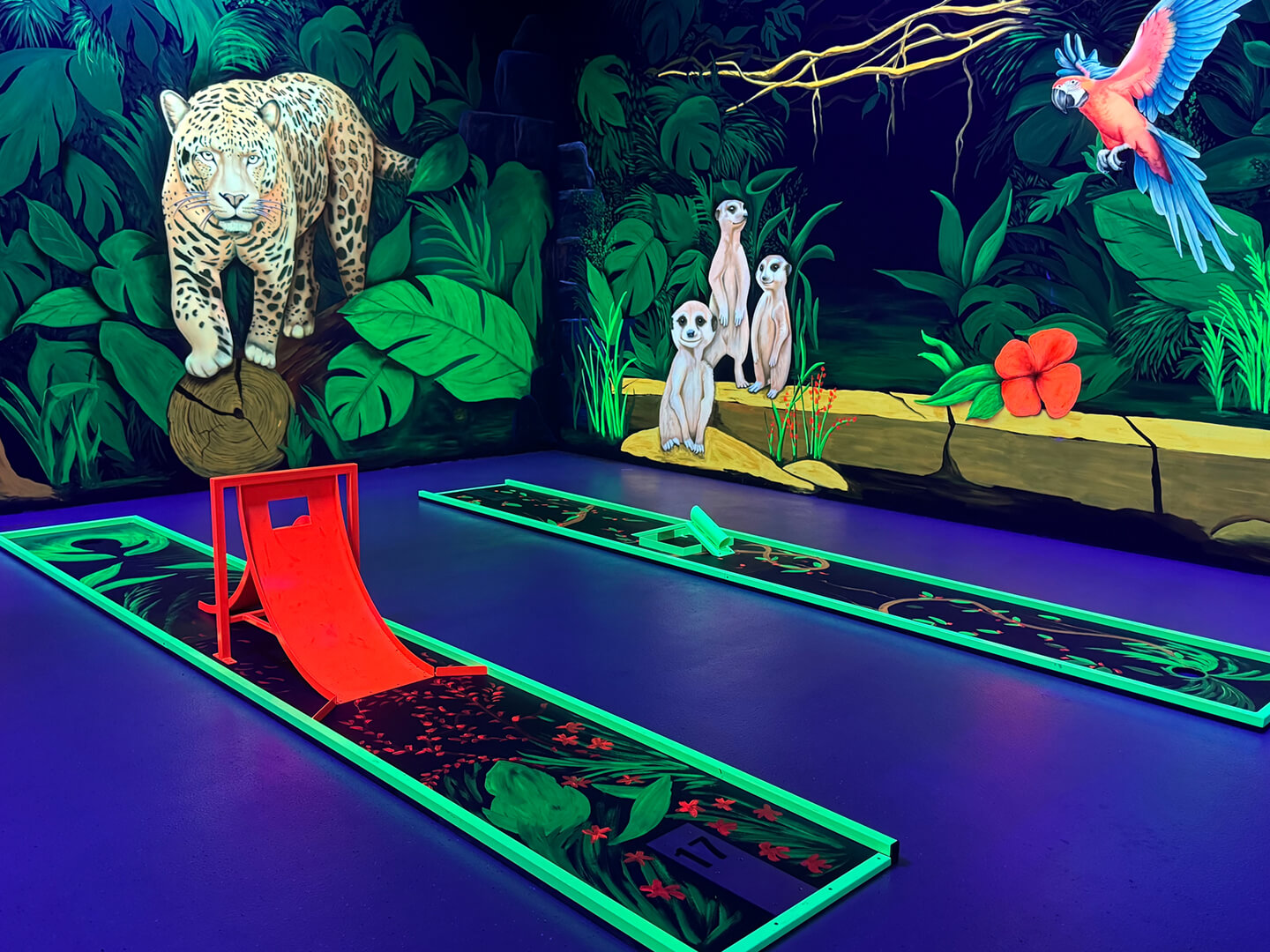 Ein weiteres Foto in die Minigolfhalle. Bunte Bilder von Tieren wie Leoparden und Erdmännchen schmücken die Wände und sind von Schwarzlicht beleuchtet.